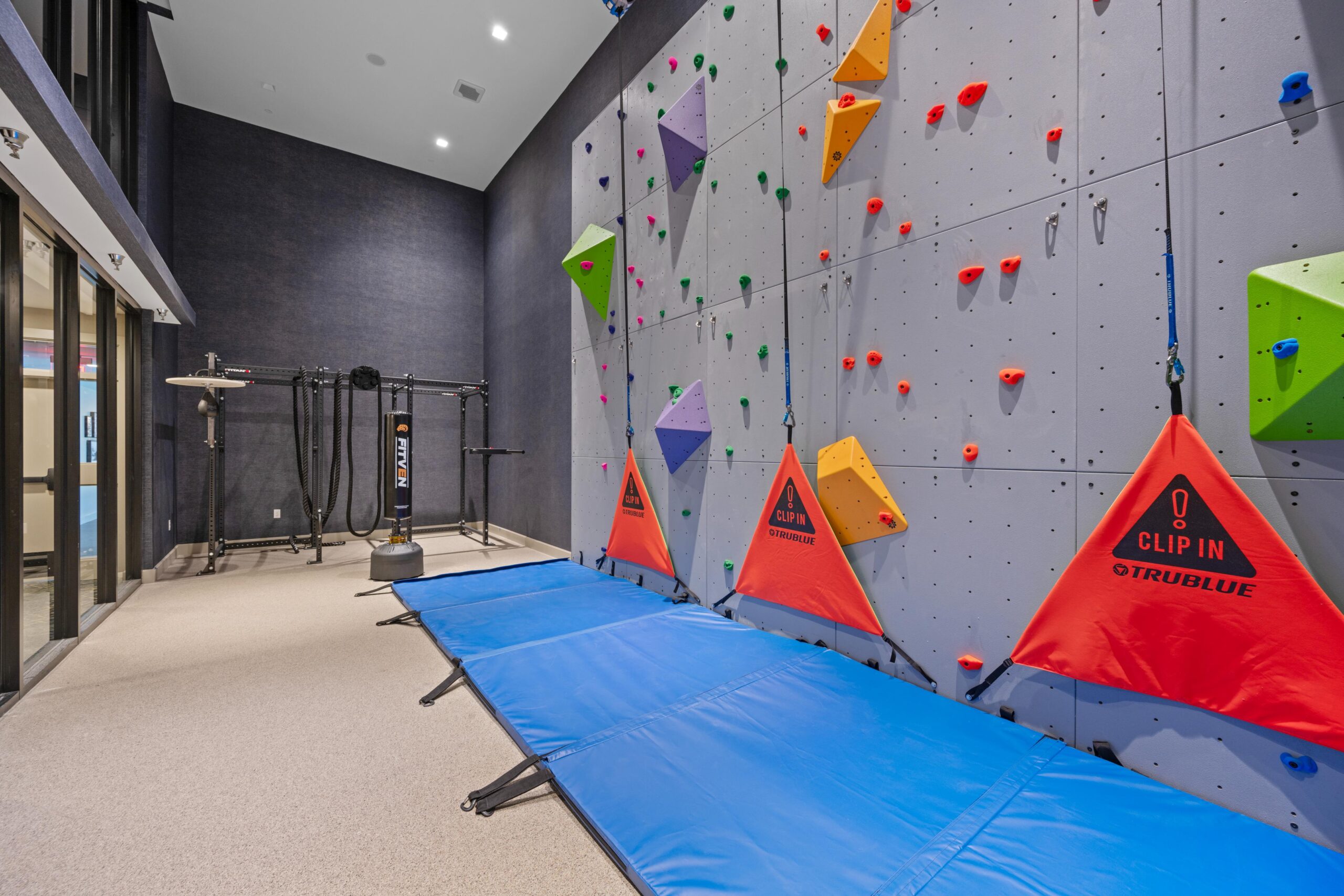 Indoor rock climbing wall with vibrant holds, safety gear, blue mats, and exercise equipment in a high-ceilinged space.