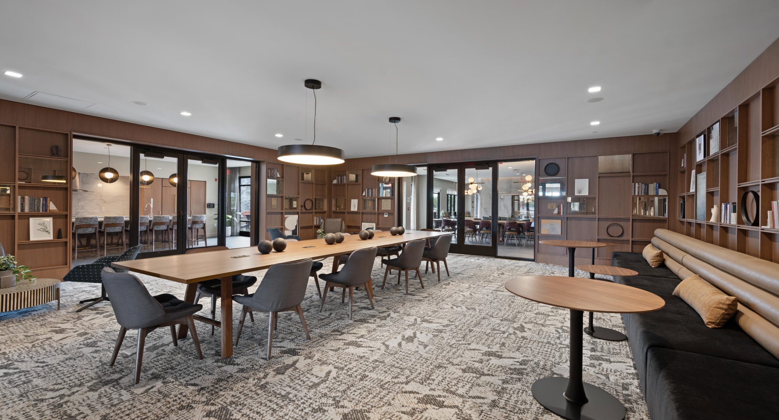 Modern meeting room with a long wooden table, grey chairs, pendant lighting, and a wall of bookshelves and glass doors.