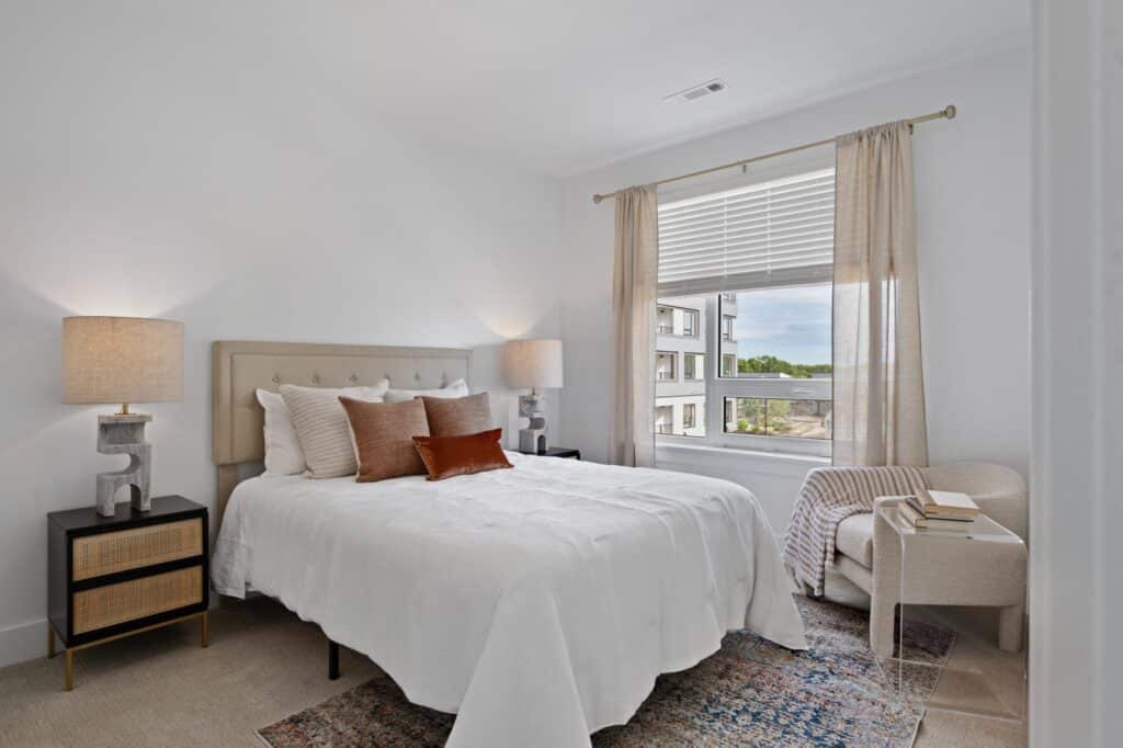 Minimalist bedroom with a white bed, beige headboard, two side lamps, a large window, and a cozy chair with books.