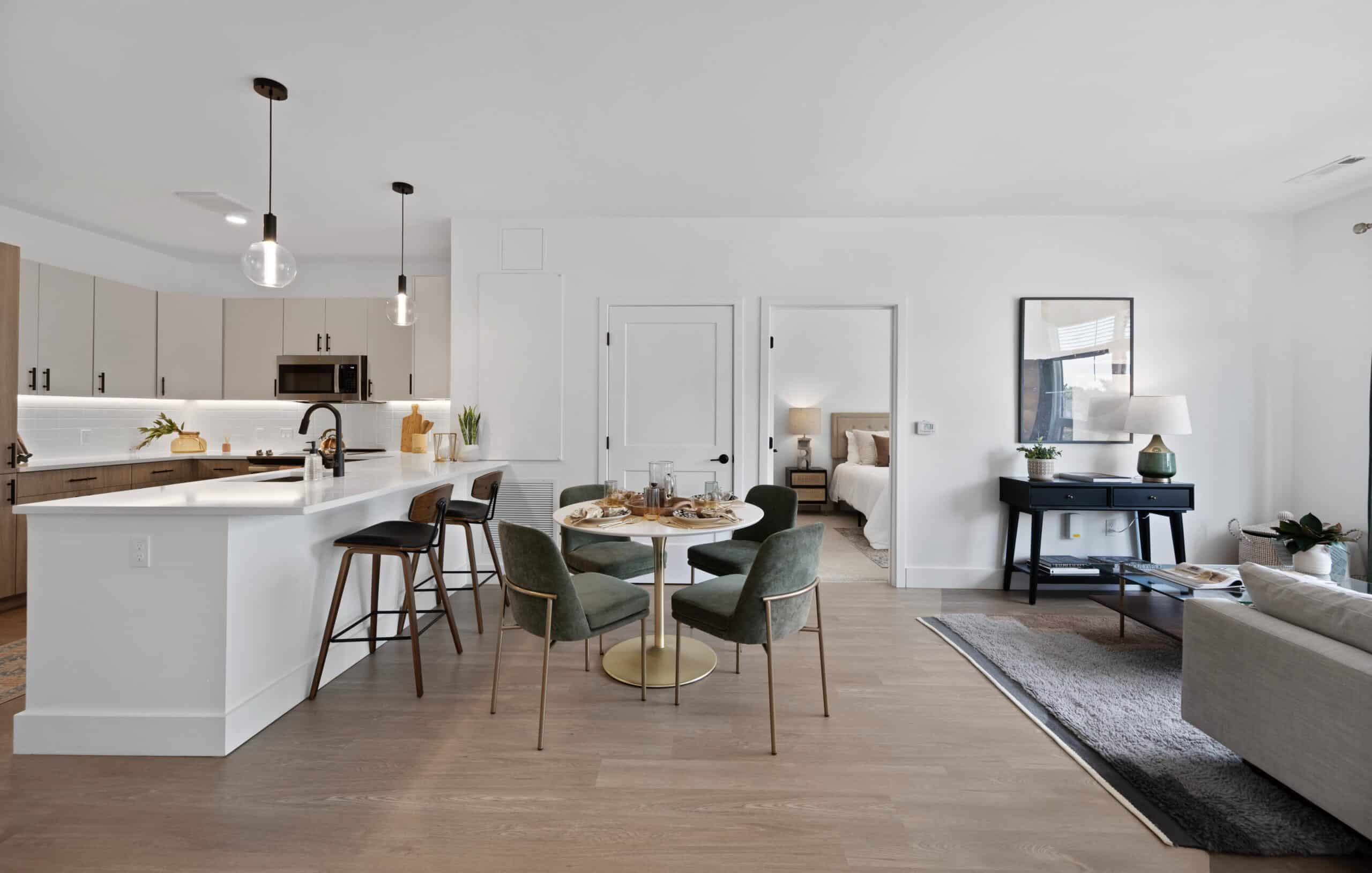 Open-concept dining and kitchen area with a round table, green chairs, bar stools at the island, and a view into a bedroom.