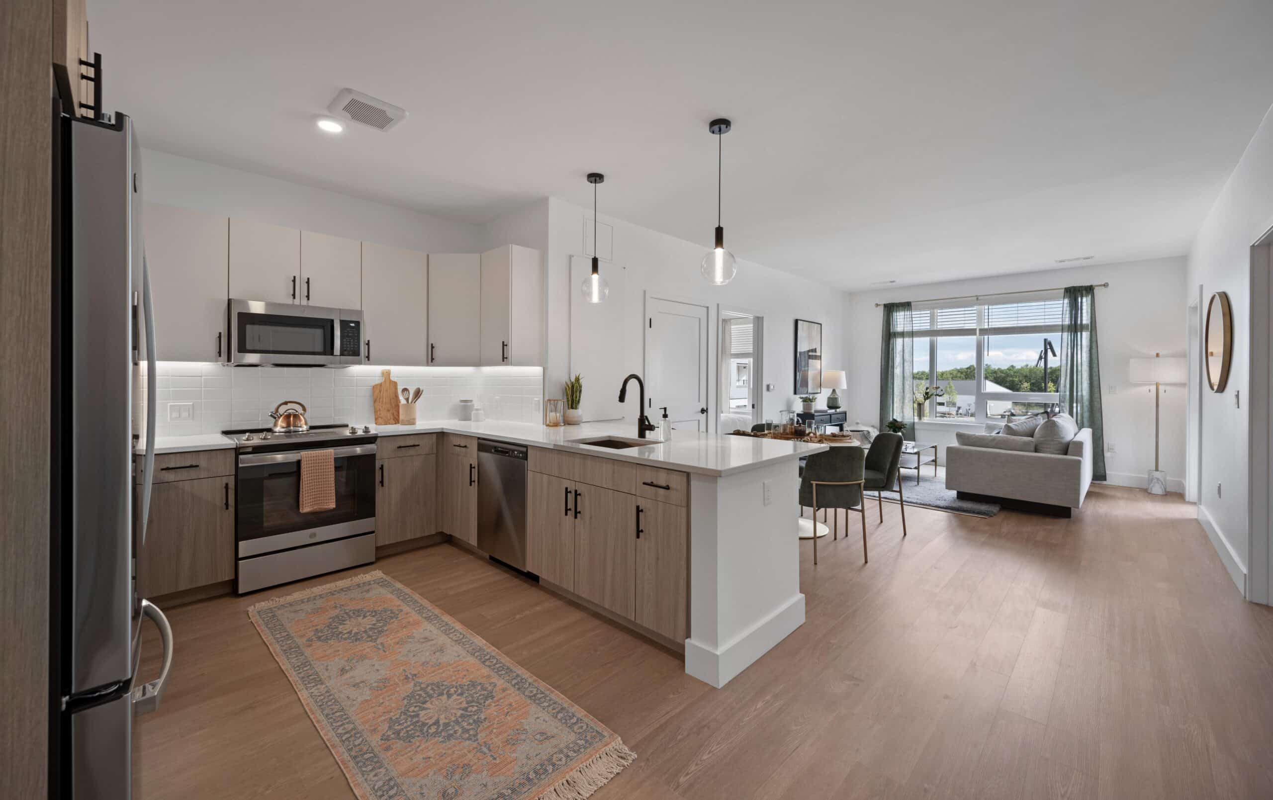 Modern kitchen with light wood cabinets, stainless steel appliances, an island, pendant lights, and open view to a bright living area.