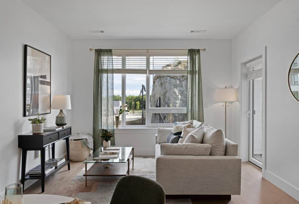 Cozy living room with a beige sofa, glass coffee table, black console, green curtains, and large window showcasing outdoor scenery.