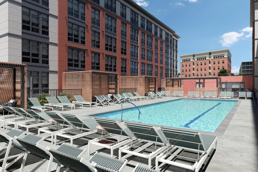 Rooftop pool deck with rows of sun loungers, cabanas, and a clear blue swimming pool set against a backdrop of red-brick buildings.