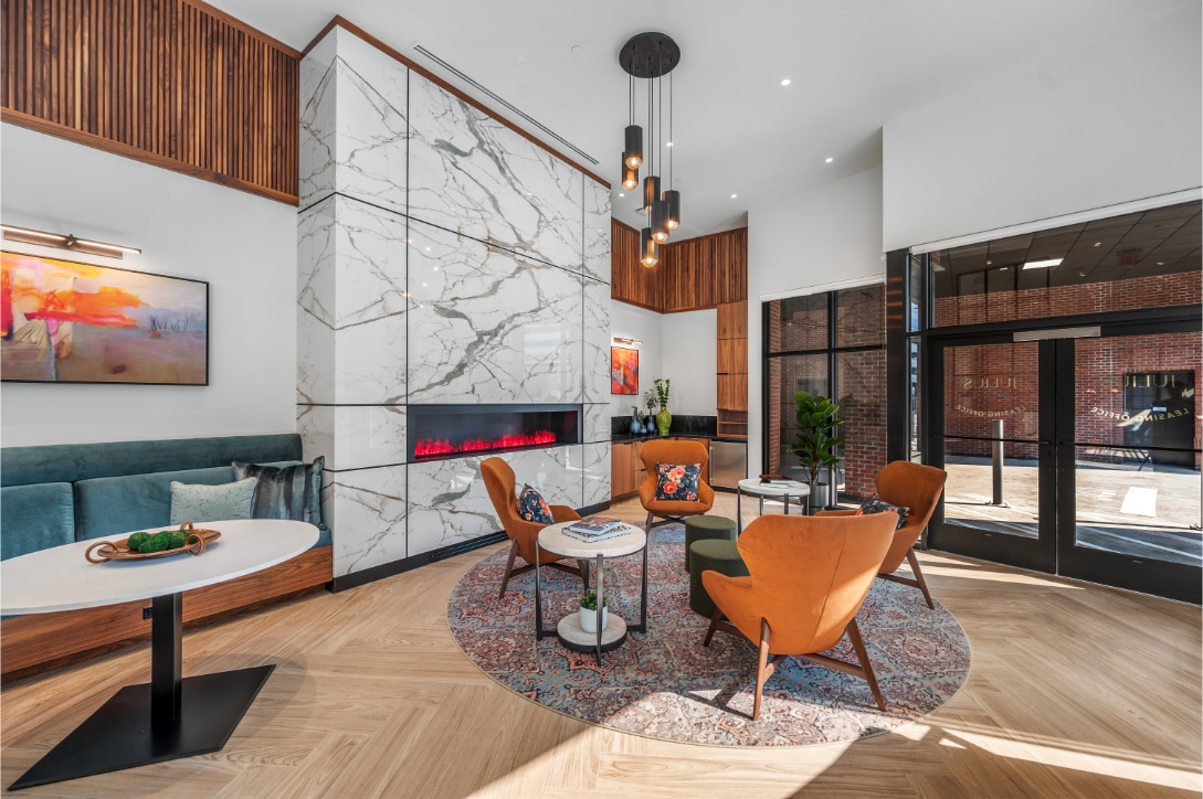 Cozy lounge with marble fireplace, warm orange chairs, a round rug, and large windows, offering a welcoming space