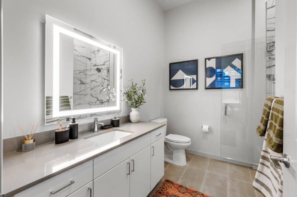 Contemporary bathroom with a spacious vanity, illuminated mirror, and geometric art, featuring a glass-enclosed shower.