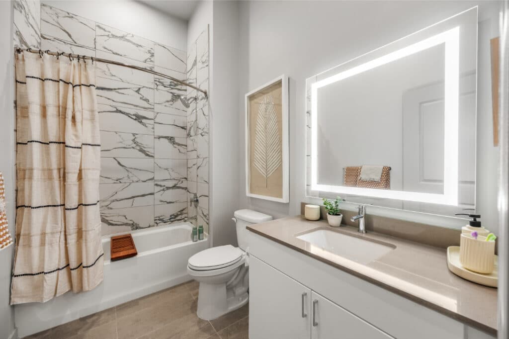 Modern bathroom with a lighted mirror, marble shower tiles, and a beige countertop, accented by minimal decor.