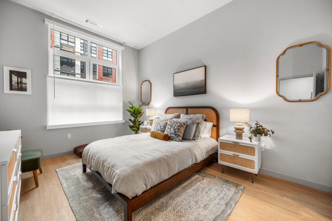 Cozy bedroom with a rattan headboard, layered bedding, matching nightstands, and a large window with a modern urban view.