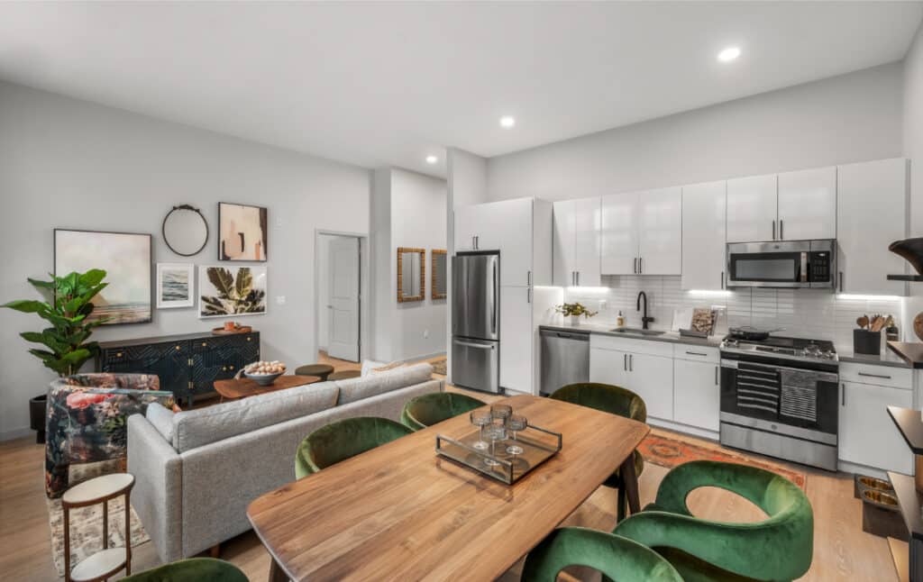 Open-plan kitchen and living area with green velvet chairs, a wood dining table, and modern white cabinetry with stainless steel appliances.