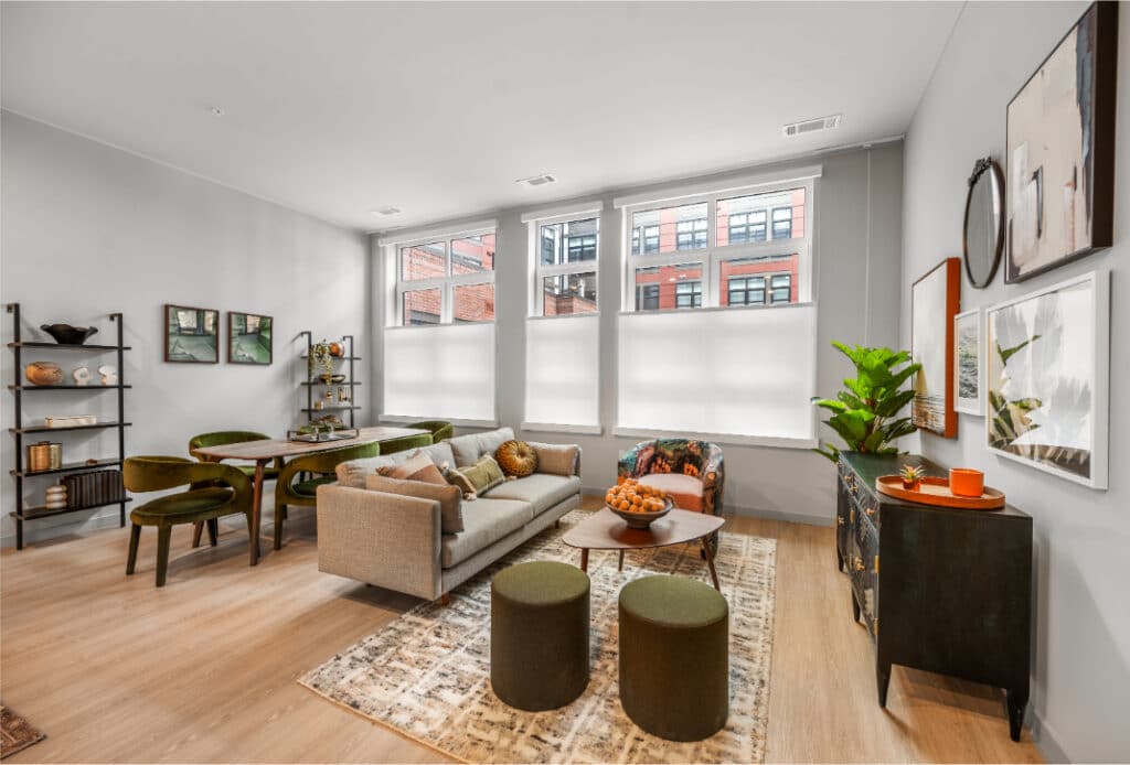 Bright living and dining space with a neutral sofa, green dining chairs, wall art, and large windows with shades.