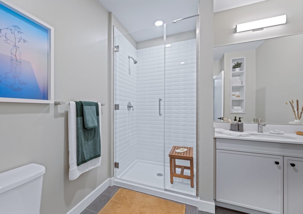 Minimal bathroom with a glass-enclosed shower, textured white tiles, and a wood bench, accented by a vibrant wall print.