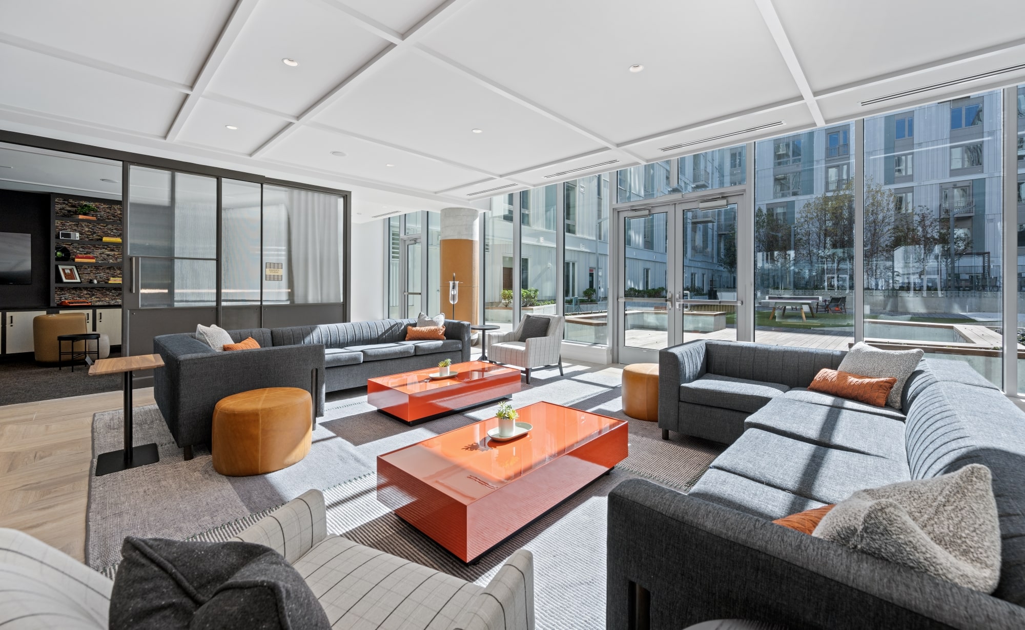 Lounge area with floor-to-ceiling windows, gray seating, and bold orange coffee tables, overlooking an outdoor courtyard.