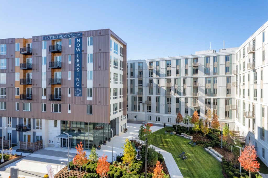 Aerial view of a modern apartment complex with landscaped gardens and a "Now Leasing" banner on the exterior.