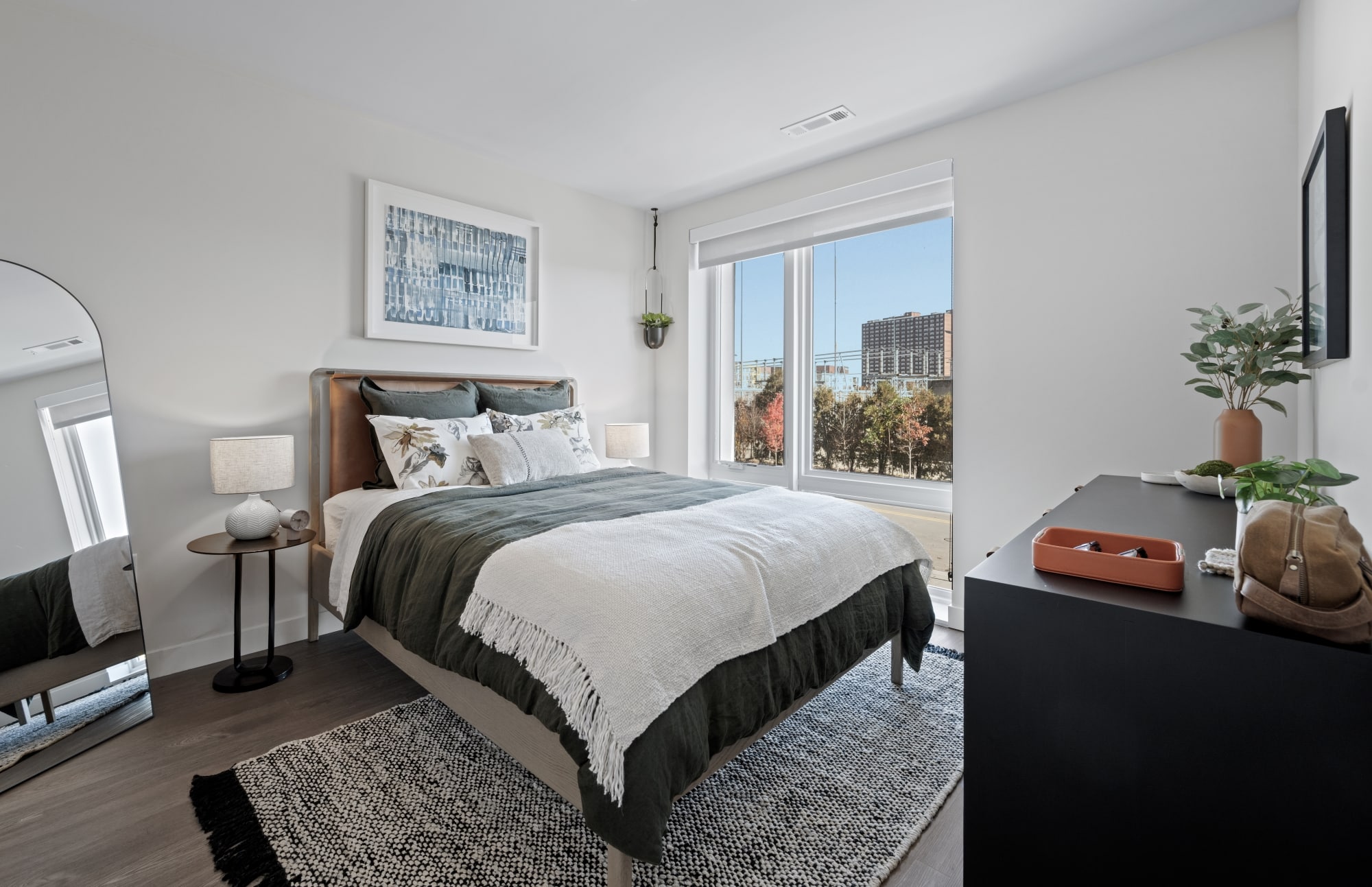 Bedroom with a large window, neutral bedding, a textured rug, and decor accents like a wall print and indoor plant.