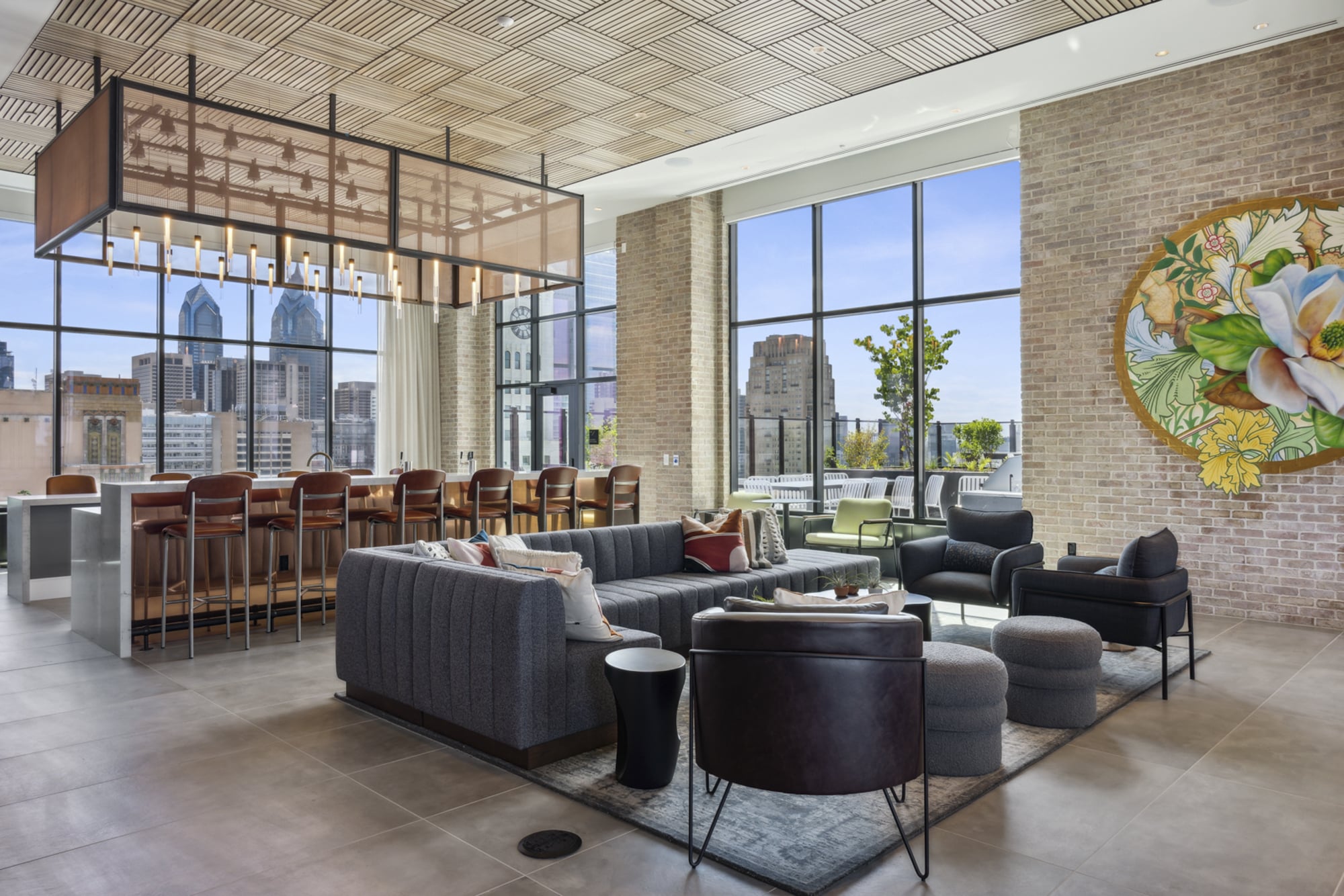 Rooftop lounge at Broad + Noble with bar, seating area, brick wall with mural, and large windows with city skyline views