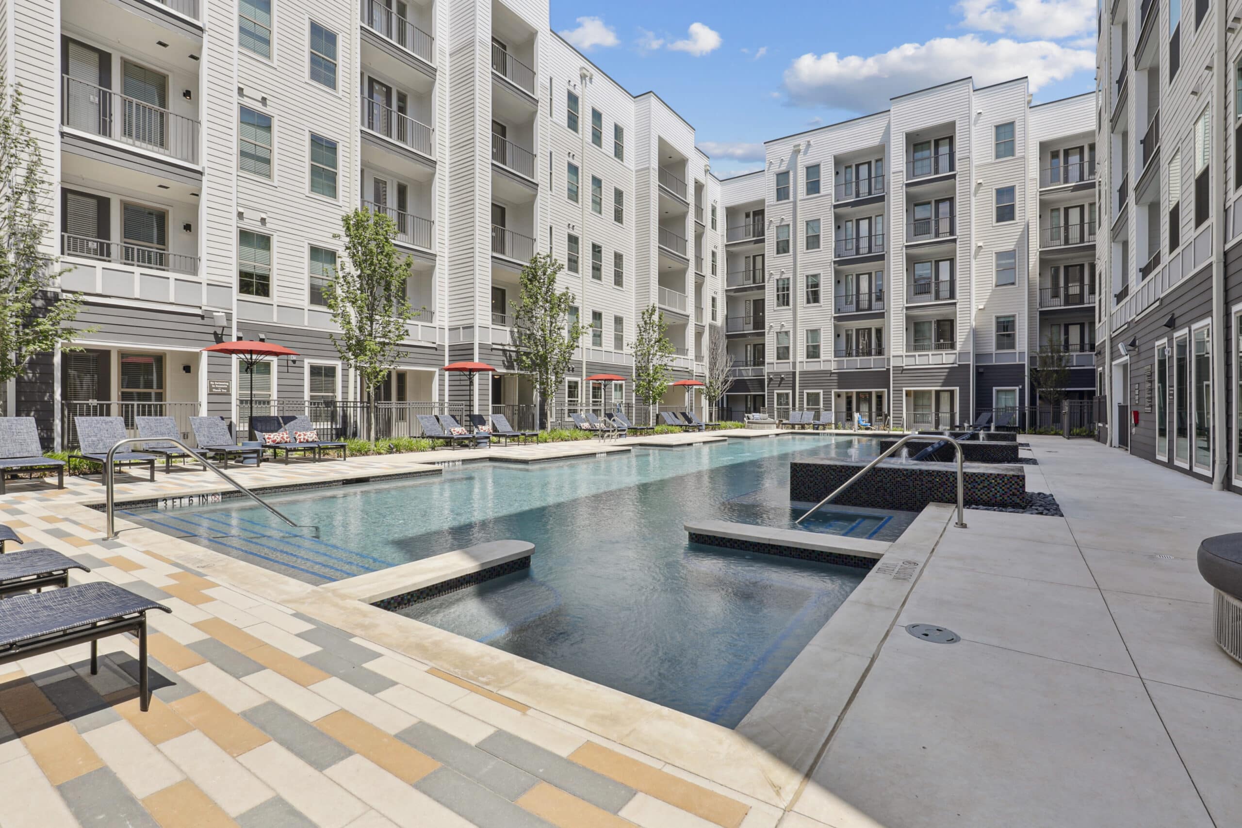 Outdoor pool area surrounded by multi-story apartment buildings, lounge chairs, and small trees.